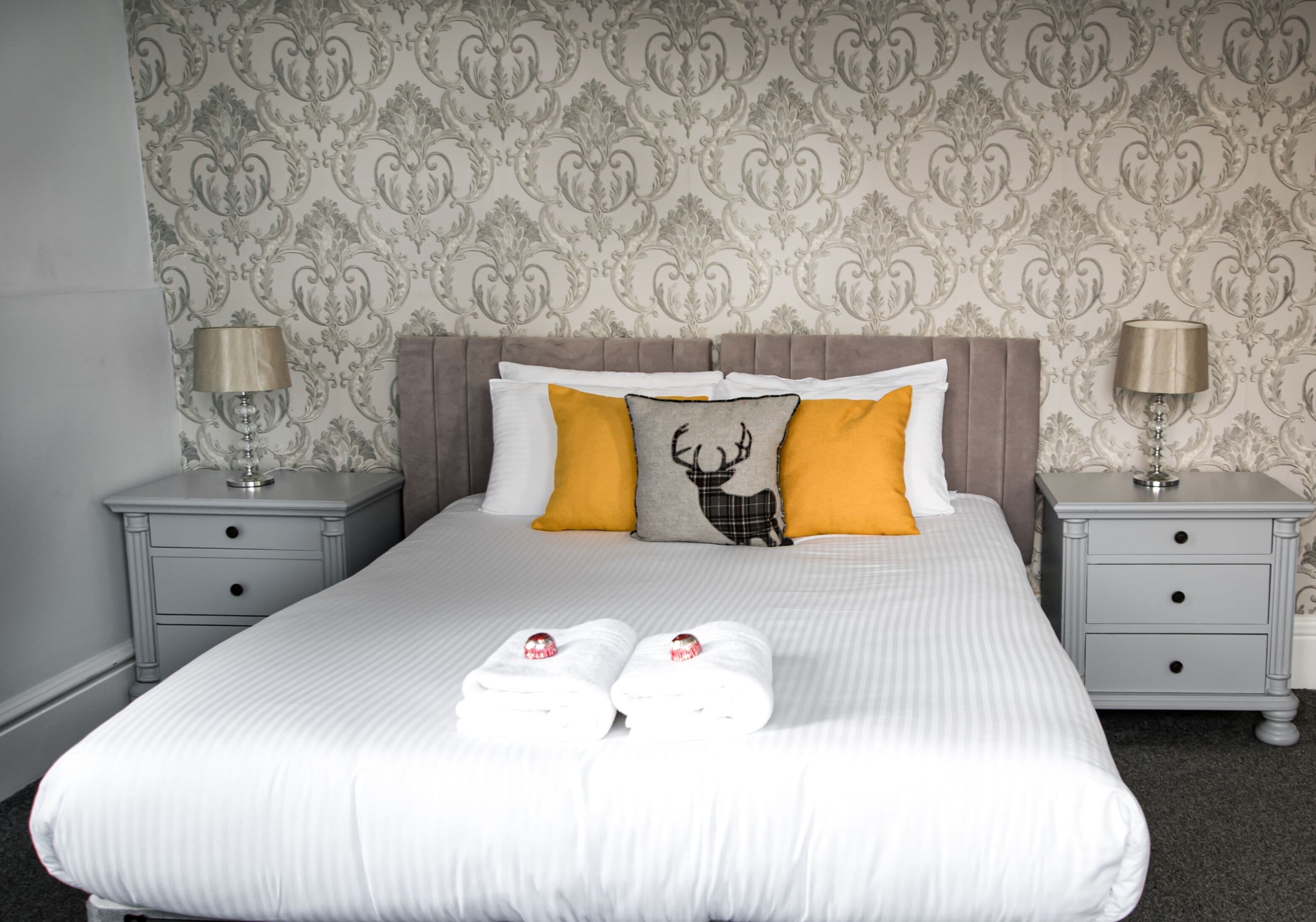 Un lit bien fait, avec du linge de lit blanc, deux oreillers et un coussin décoratif orné d’un motif de cerf. Il y a deux tables de chevet avec des lampes et un petit tiroir.