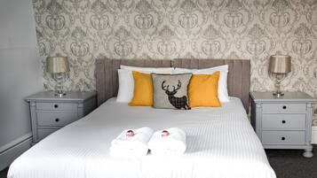 Un lit bien fait, avec du linge de lit blanc, deux oreillers et un coussin décoratif orné d’un motif de cerf. Il y a deux tables de chevet avec des lampes et un petit tiroir.