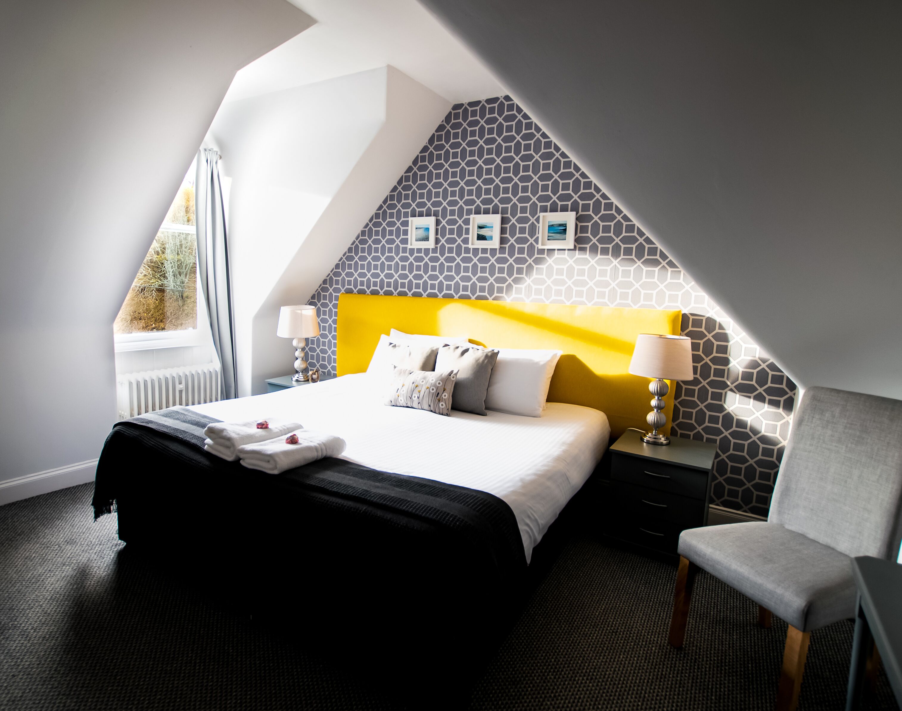 Una habitación de hotel moderna con una cama, dos mesitas de noche, una silla y un ventanal con cortinas.