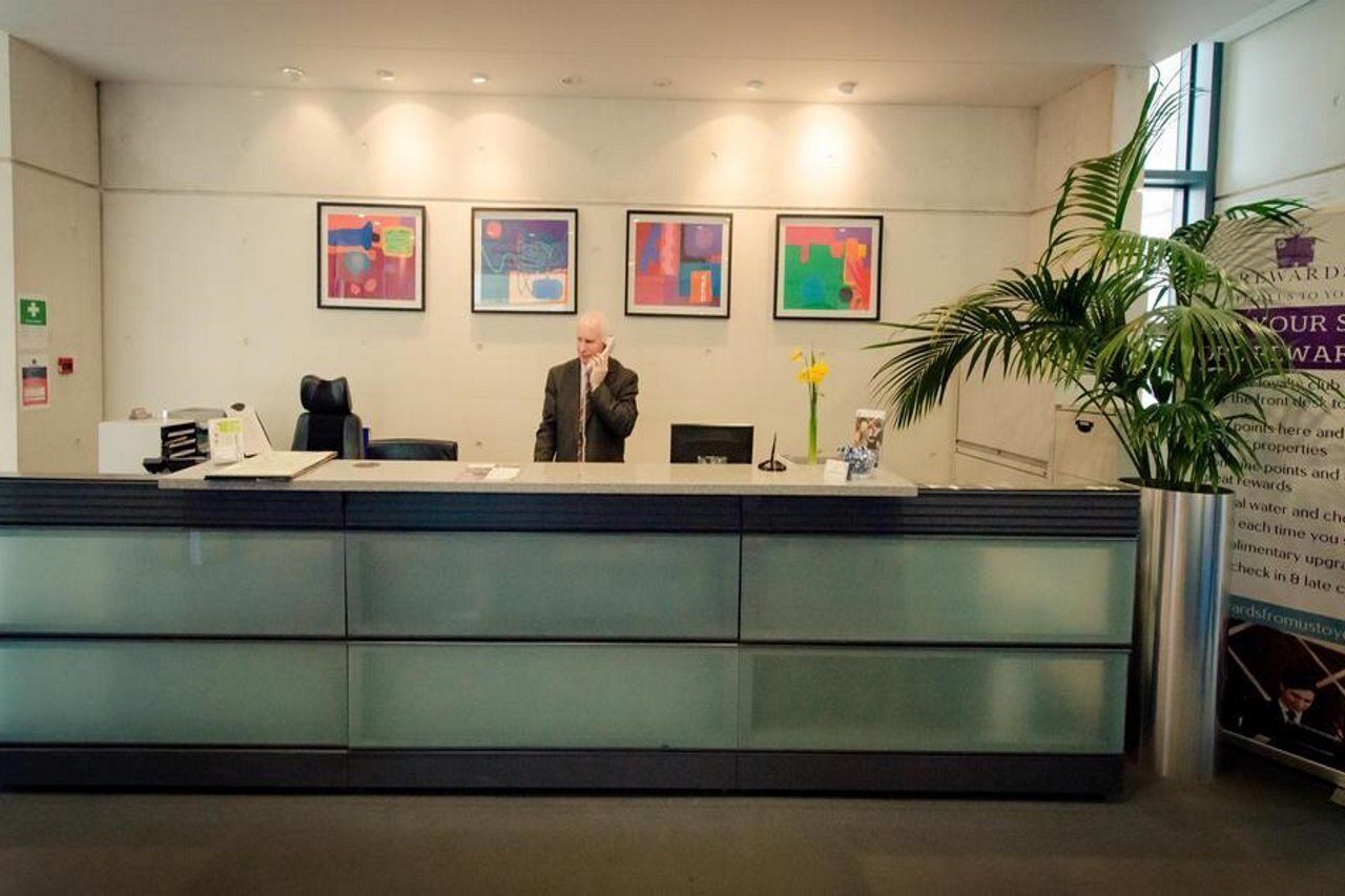 A reception desk with a man standing behind it, framed abstract art on the wall, and a large potted plant to the right.