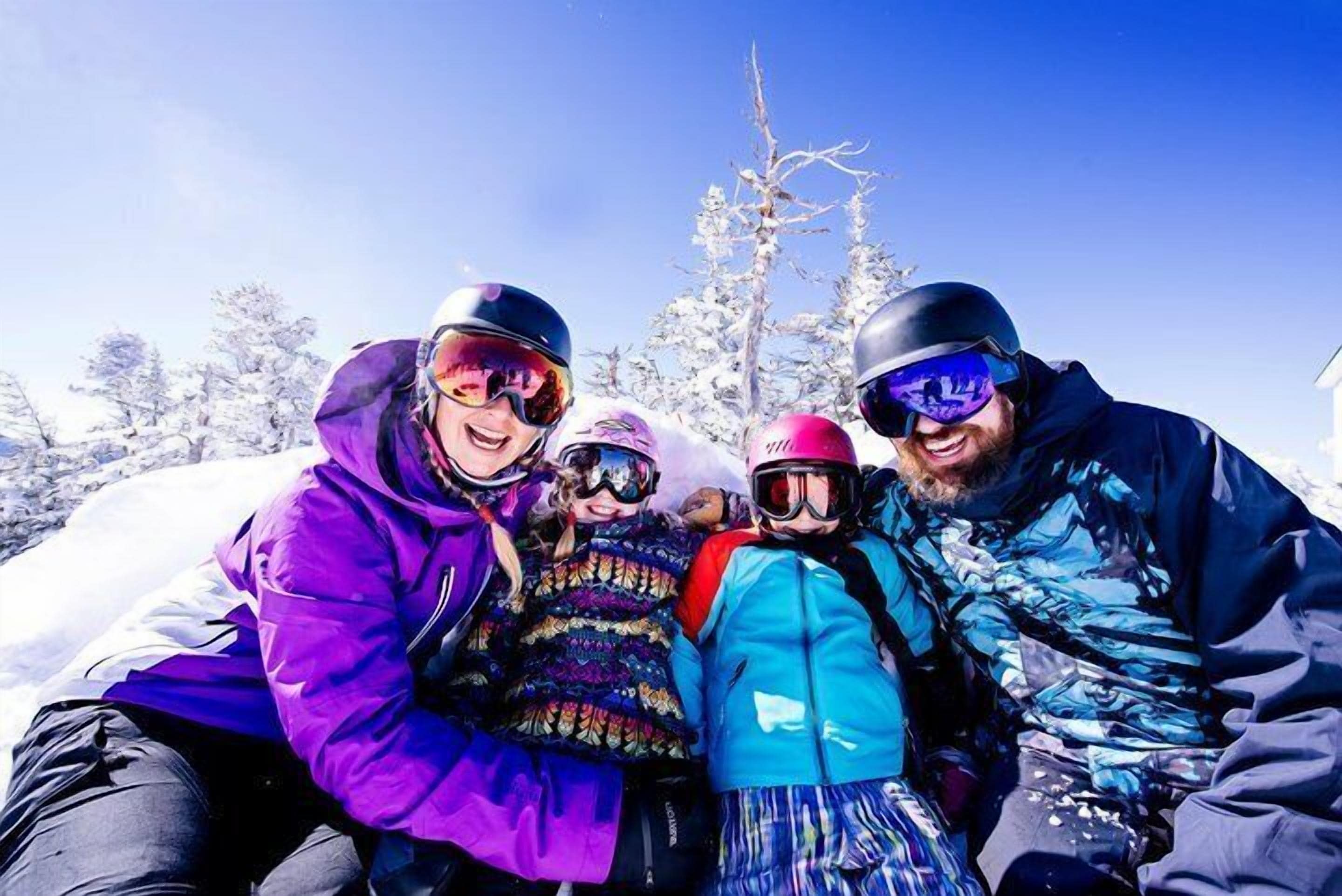 A group of people in winter clothing and helmets, sitting on a sled in a snowy landscape with trees in the background.