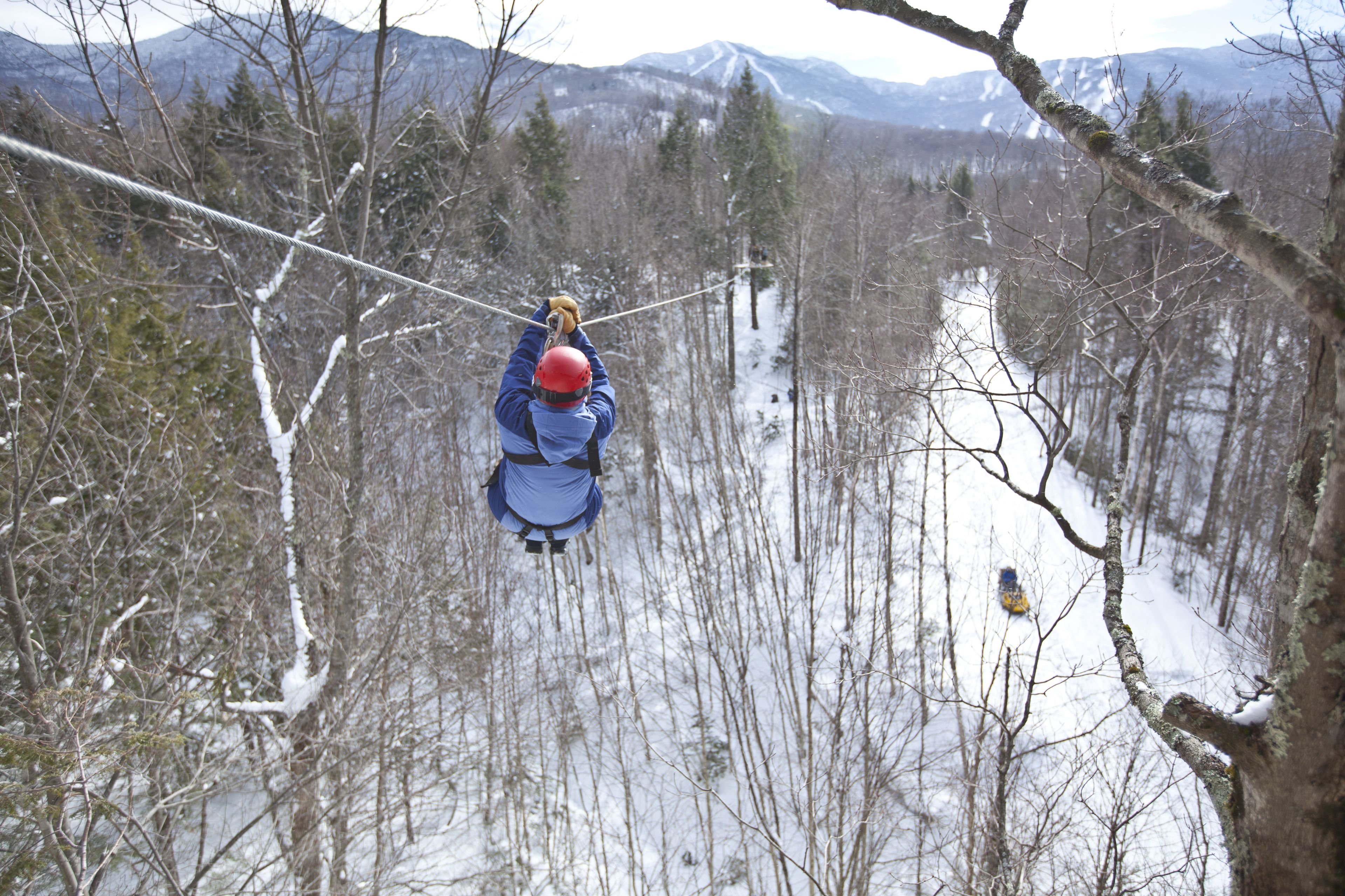 Une personne qui fait de la tyrolienne à travers une forêt enneigée, avec des montagnes en arrière-plan.
