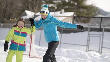 Two people in snow gear, one holding the other, in a snowy outdoor setting with a fence and trees in the background.