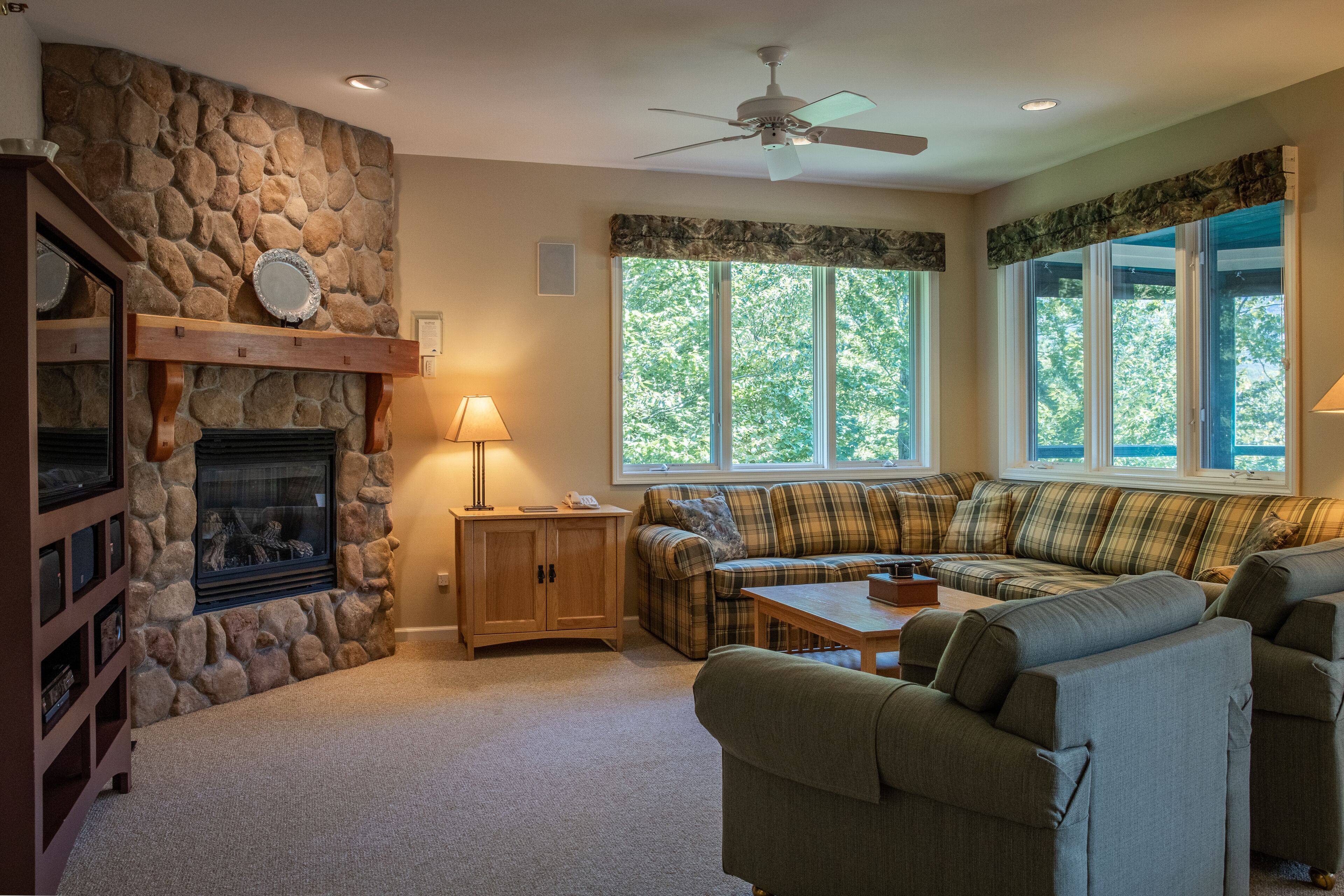 A cozy living room with a stone fireplace, a ceiling fan, and a large window with a view of trees.
