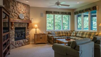 A cozy living room with a stone fireplace, a ceiling fan, and a large window with a view of trees.