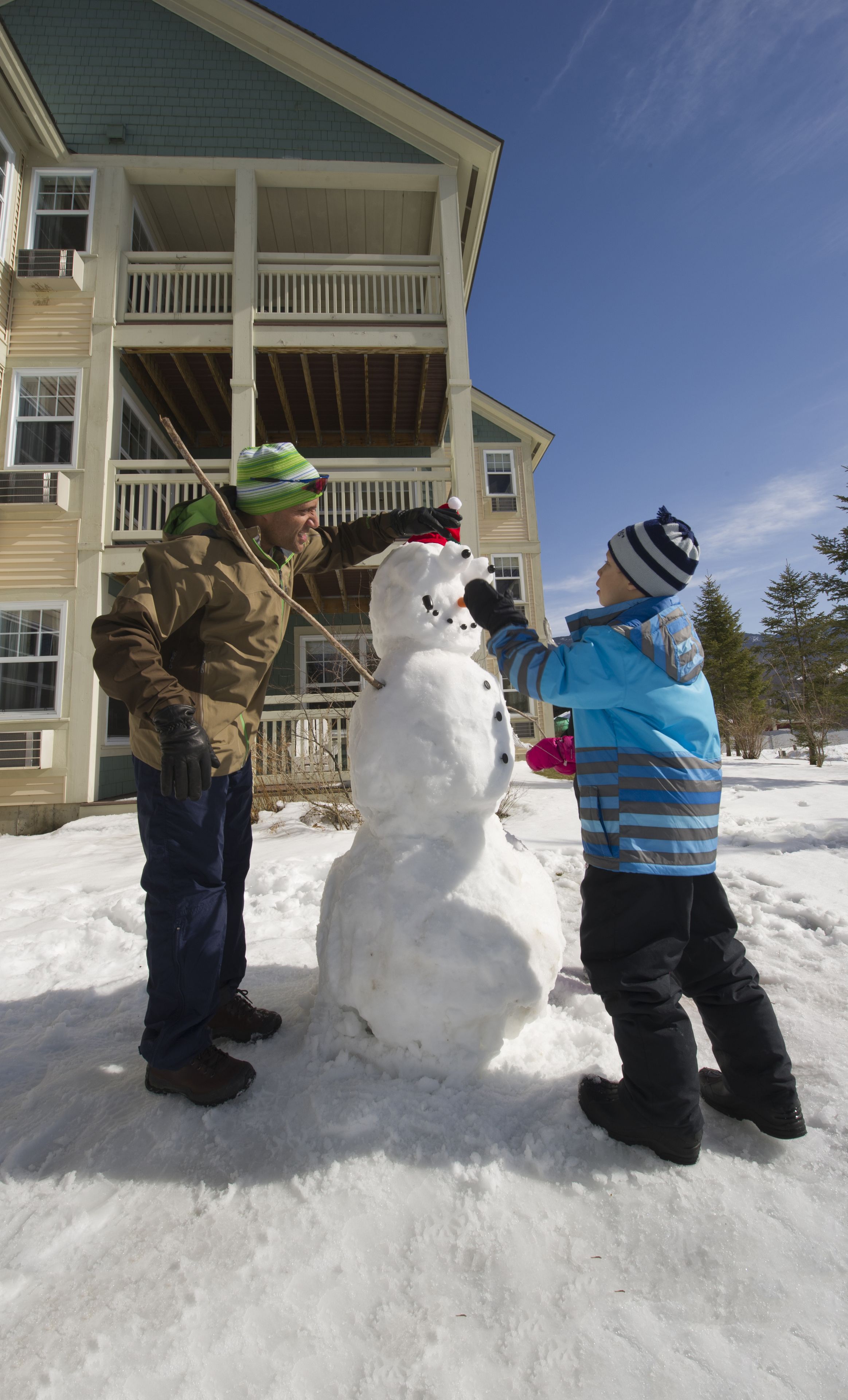 A snowman being built in front of a two-story house with a porch.