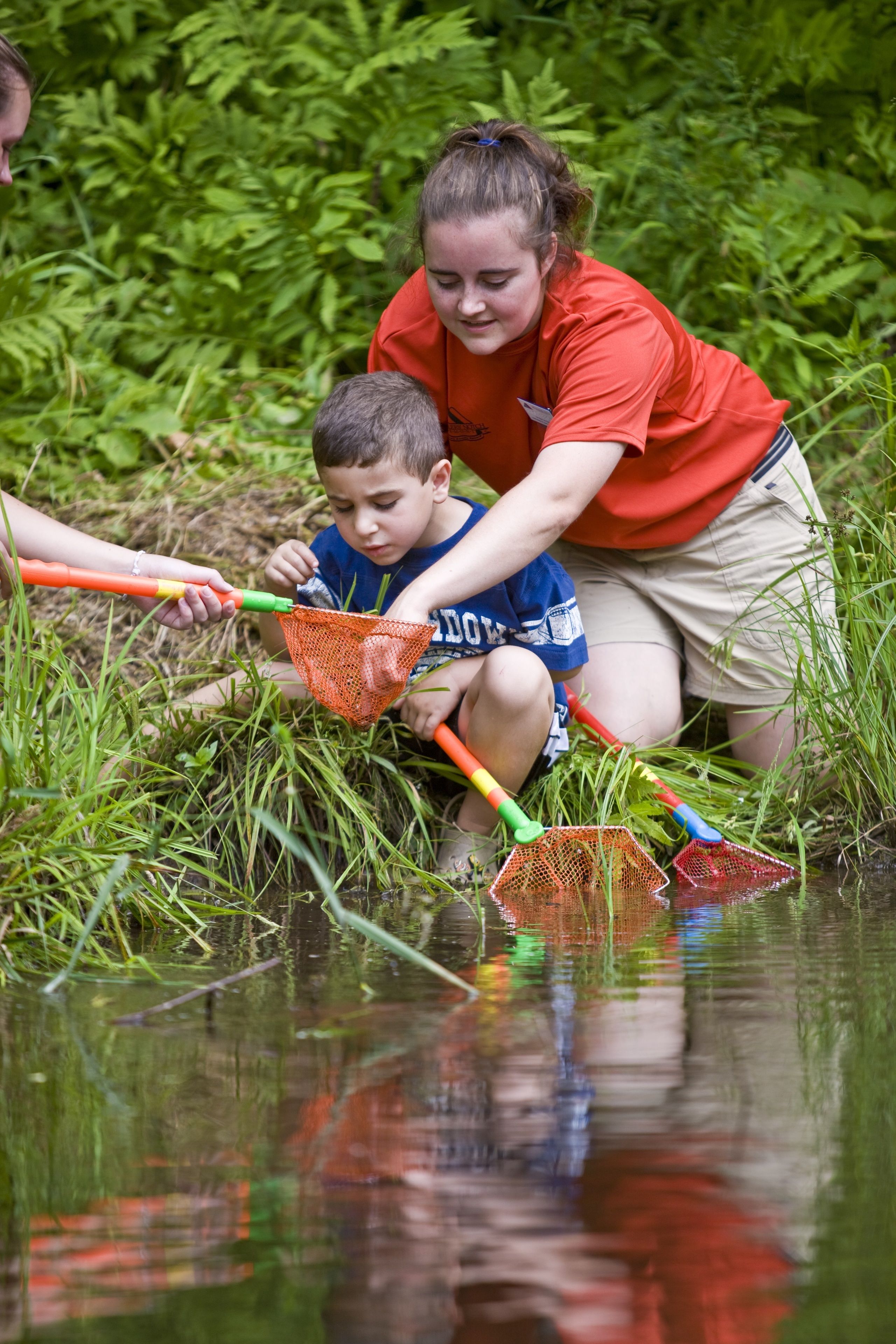 A person and a child by a water body, possibly a pond or stream, with vegetation around.