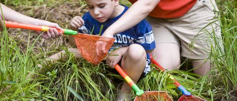 A person and a child by a water body, possibly a pond or stream, with vegetation around.