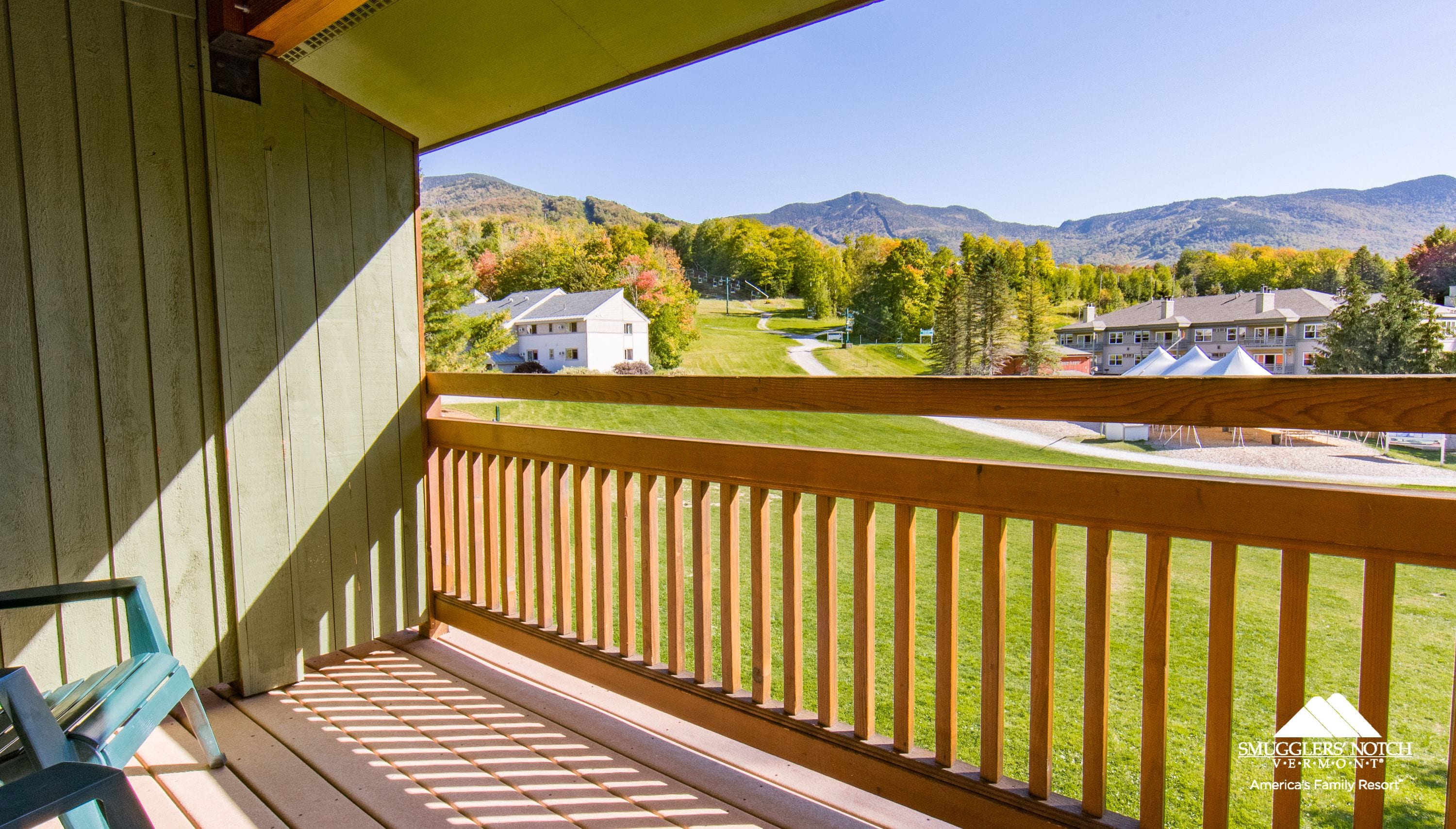 A wooden balcony with a view of a green lawn, houses, and mountains.