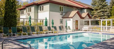 A swimming pool with lounge chairs and umbrellas, a house with a red roof, and a large evergreen tree.