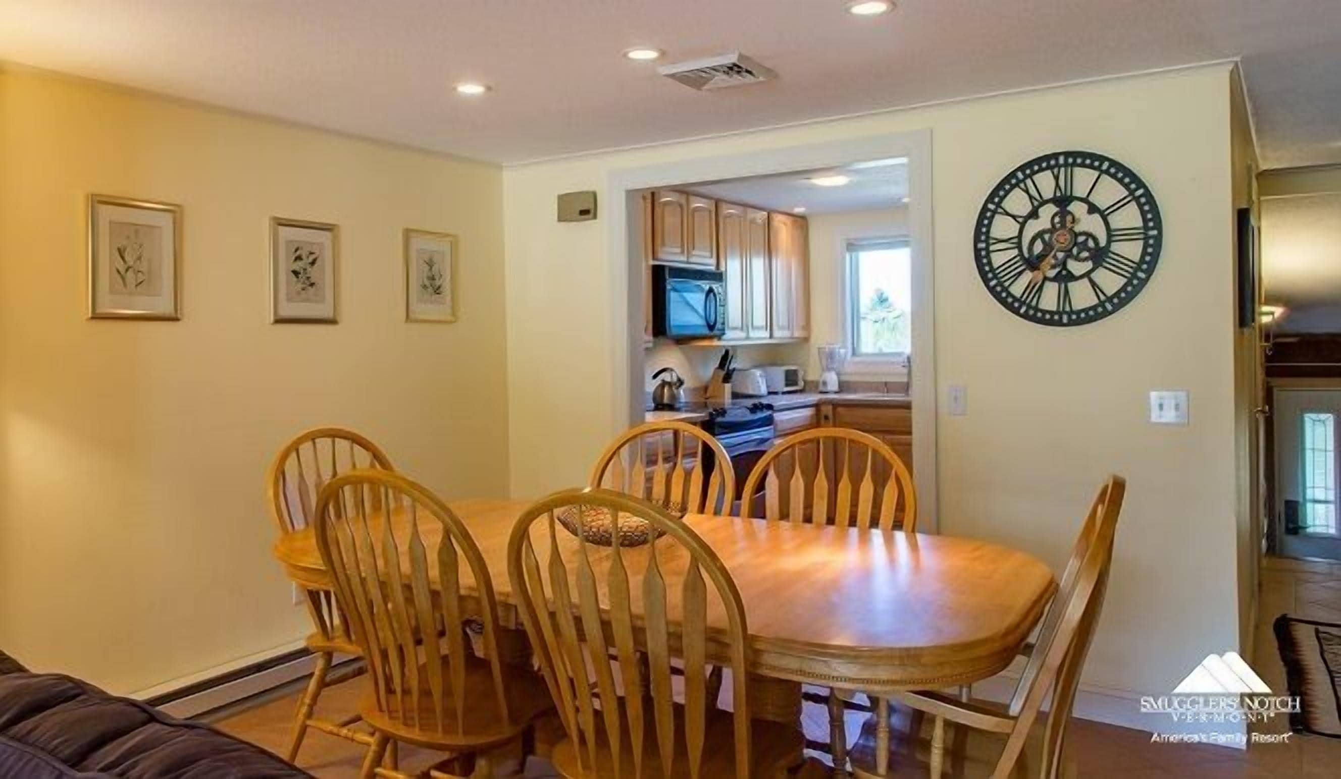 A dining area with a wooden table and chairs, a kitchen in the background, and a wall clock.