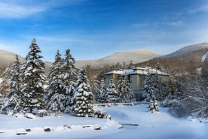 Paisaje nevado con un edificio rodeado de árboles cubiertos de nieve y montañas al fondo.