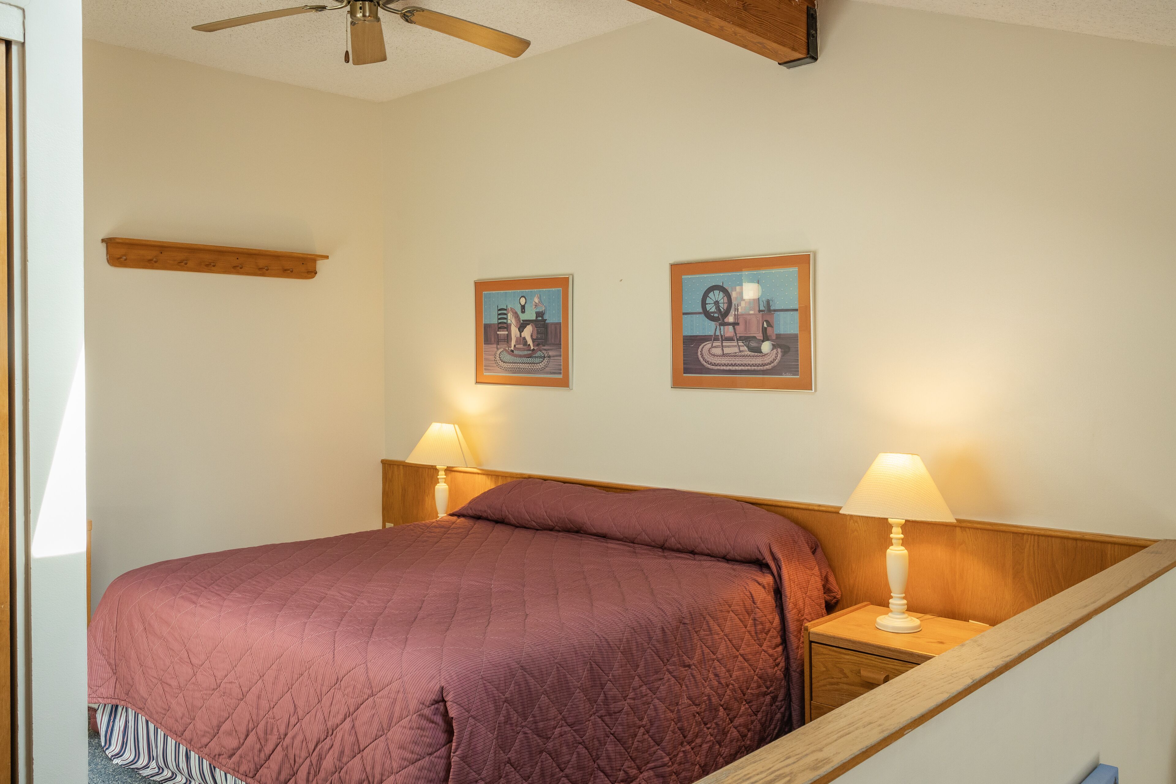 A bedroom with a bed, two bedside lamps, a ceiling fan, and two framed pictures on the wall.