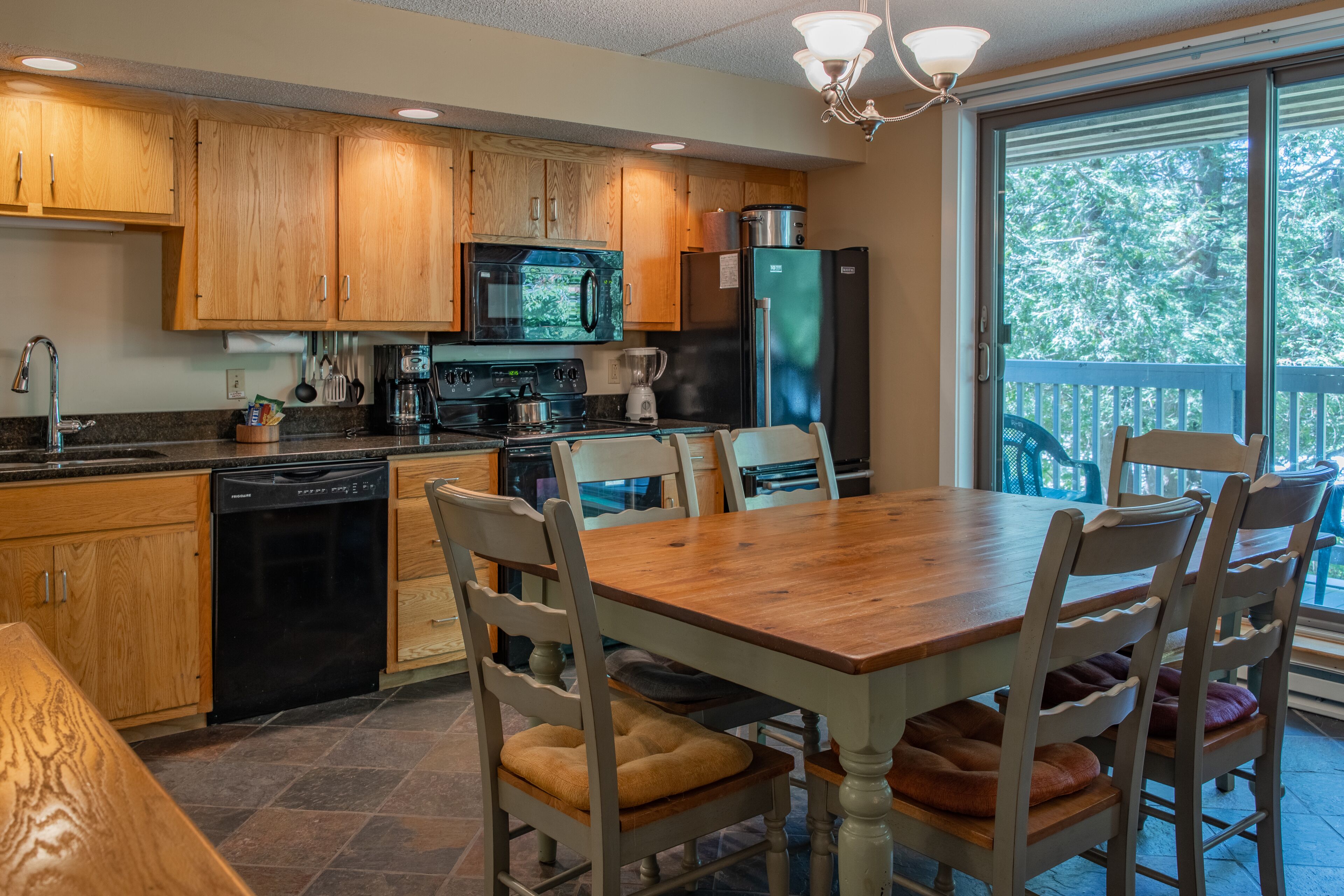 A kitchen with wooden cabinets, a microwave, stove, and refrigerator. There is a dining table with chairs and a view of the outdoors through a sliding glass door.
