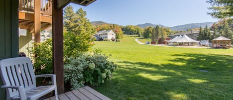 A wooden deck with a white plastic chair, a grassy lawn, and a view of a white building with a covered area in the distance.