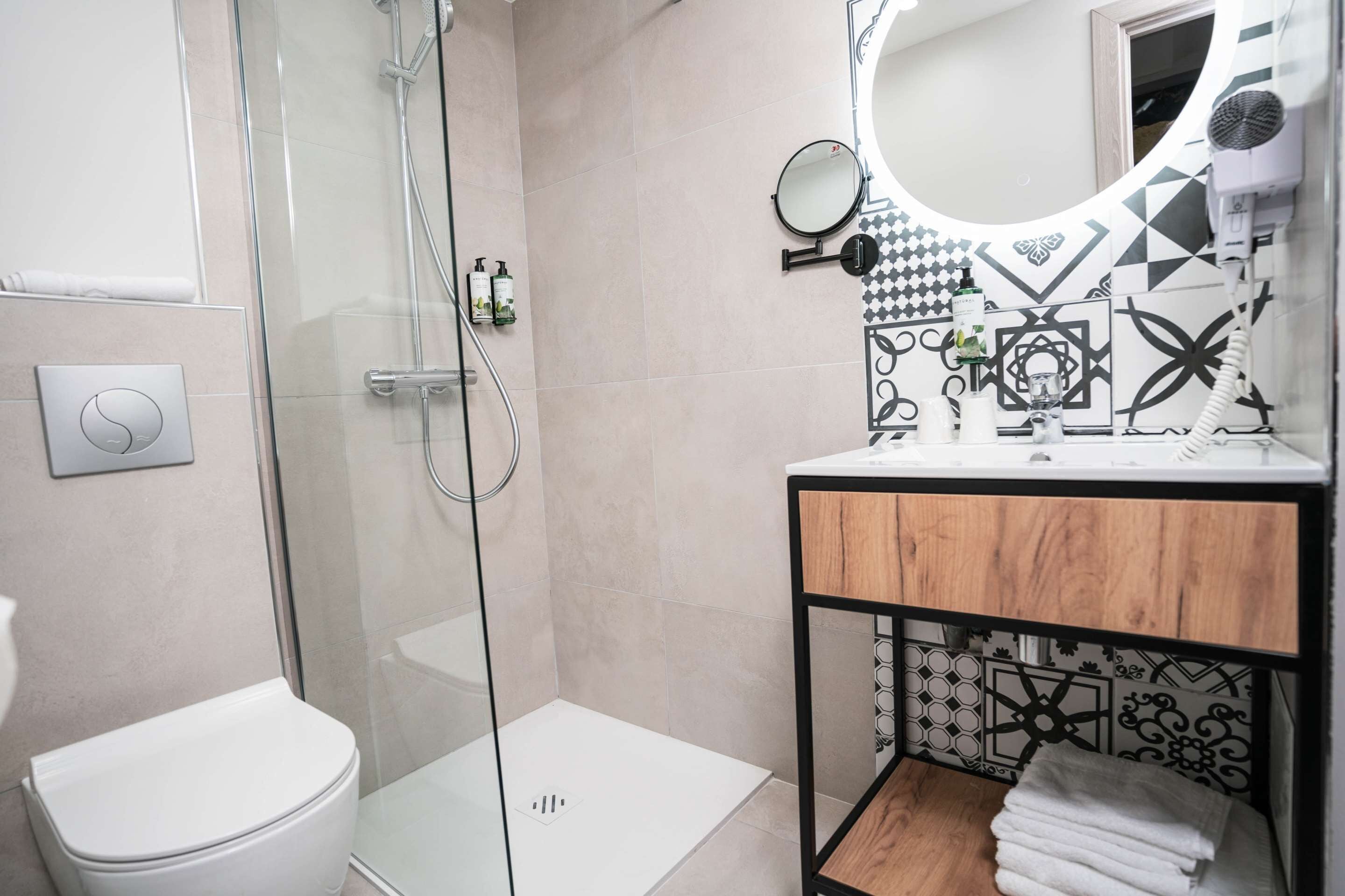 A modern bathroom with a glass shower enclosure, a toilet, a vanity with a mirror, and a shelf with towels.