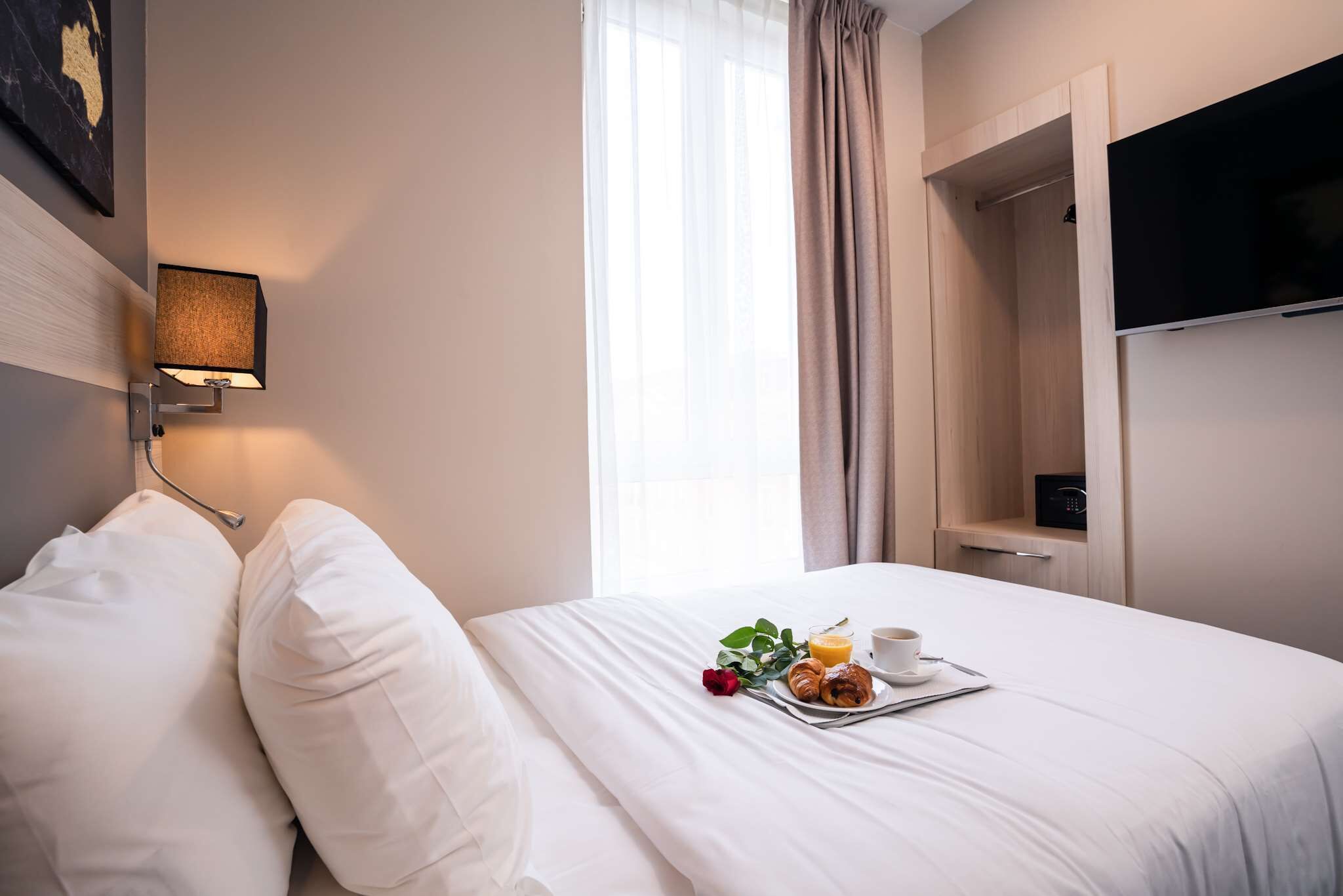 Et hotelværelse med en pænt redt seng, et sengebord med kop og kager, et fjernsyn på væggen og et vindue med gardiner.