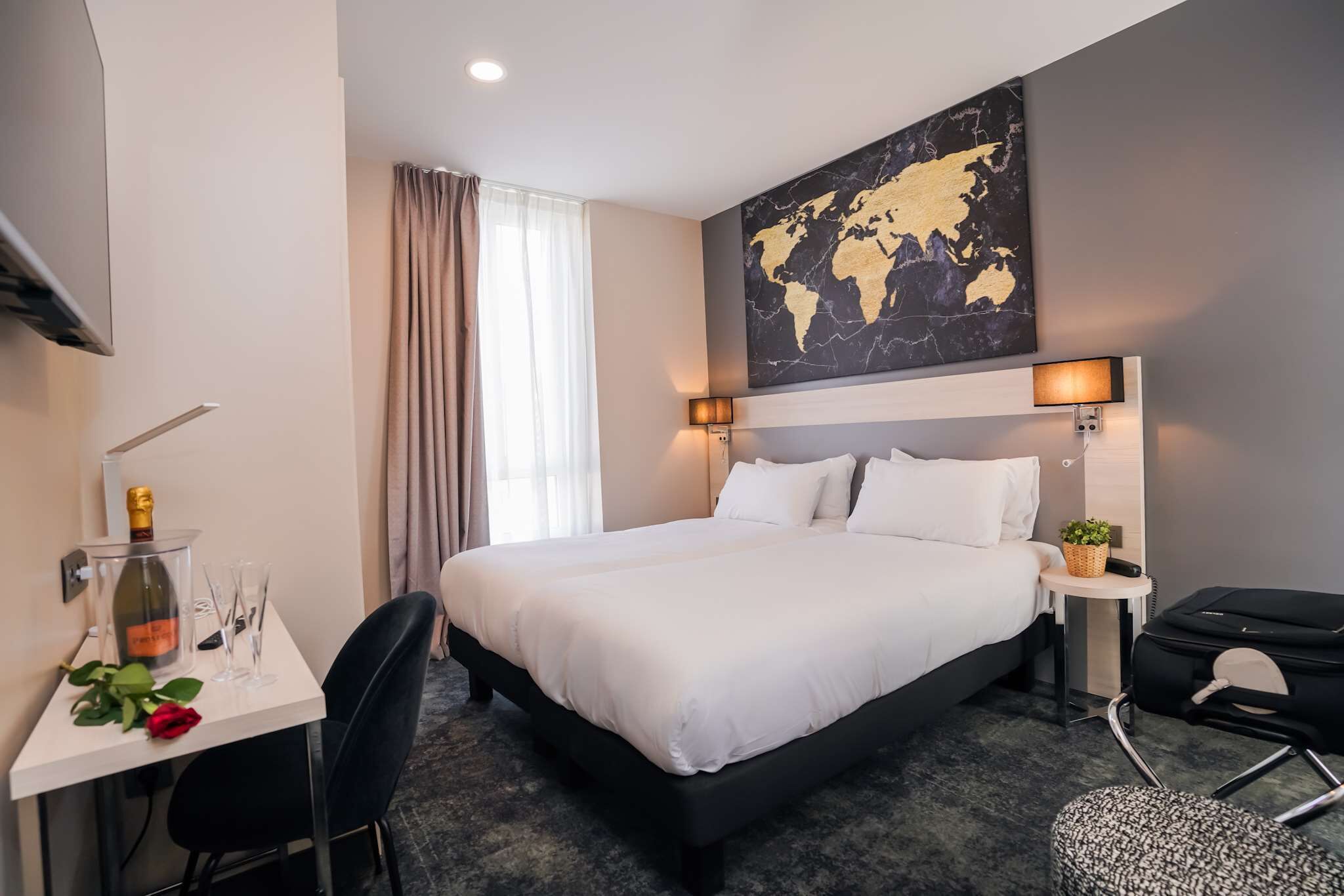 Une chambre d’hôtel avec un grand lit, un bureau sur lequel se trouve une bouteille de champagne et des verres, une carte du monde accrochée au mur et une petite table avec une plante.