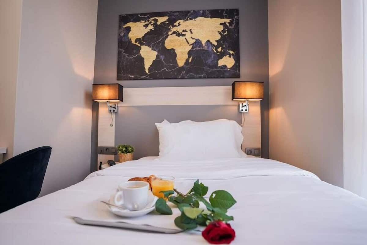 Una habitación de hotel con una cama, un mapa mundial en la pared, dos lámparas de noche, una mesita con una taza de café, un vaso de jugo de naranja y una rosa roja.