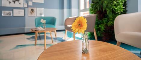 A modern hotel lobby with a round wooden table, a vase with a single flower, a blue wall with framed artwork, a green vertical garden, and a white sofa.
