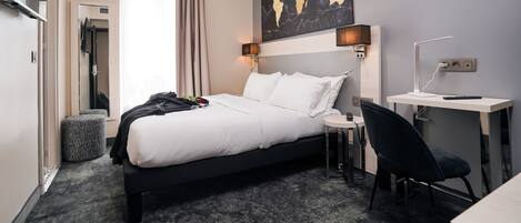 A hotel room with a bed, a desk, a chair, a world map on the wall, and a window with curtains.