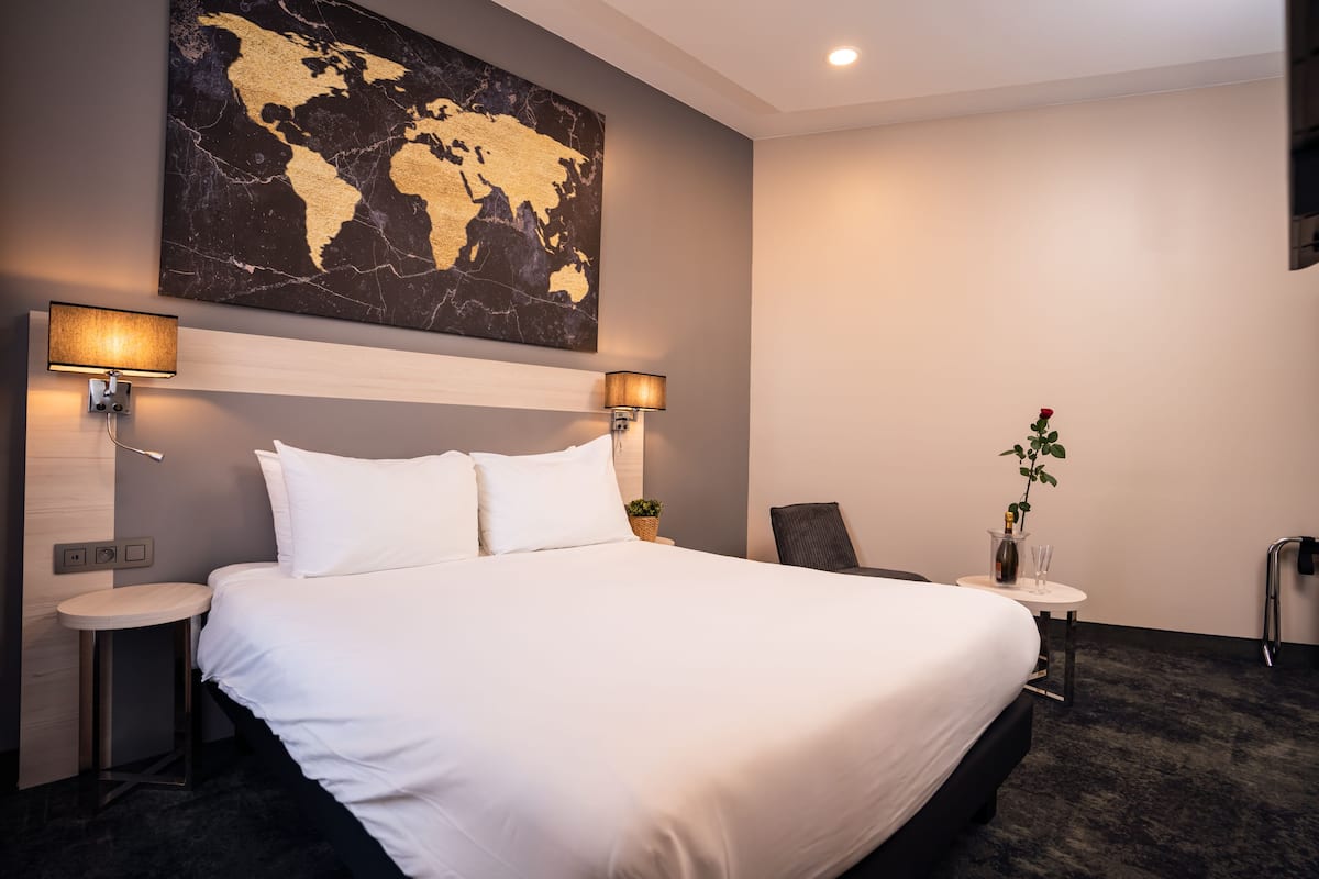 Una habitación de hotel moderna con una cama grande, dos mesitas de noche, un cuadro con un mapa mundial, una pequeña mesa con un jarrón y una planta, y una silla.