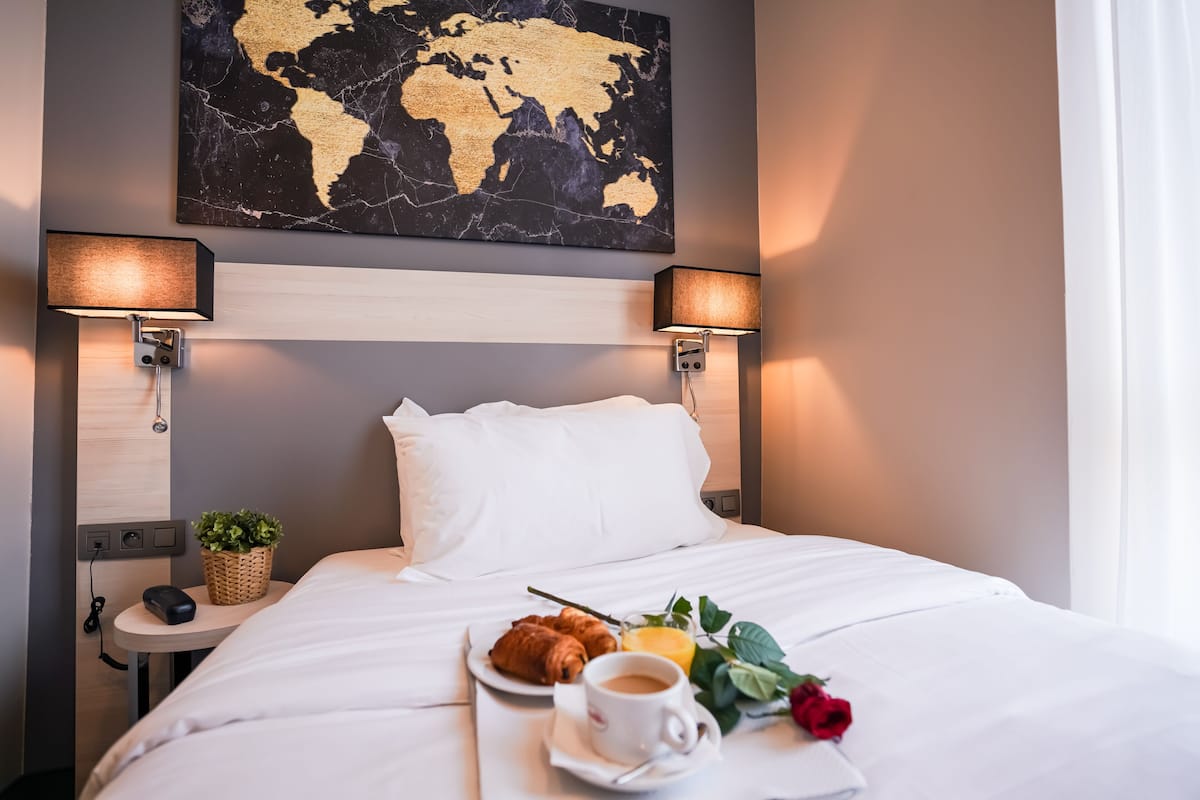 Una cama bien tendida con sábanas blancas, un cuadro de un mapa mundial encima, dos lámparas de noche, una mesita con una cesta, un plato de croissants, una taza de café y una rosa.