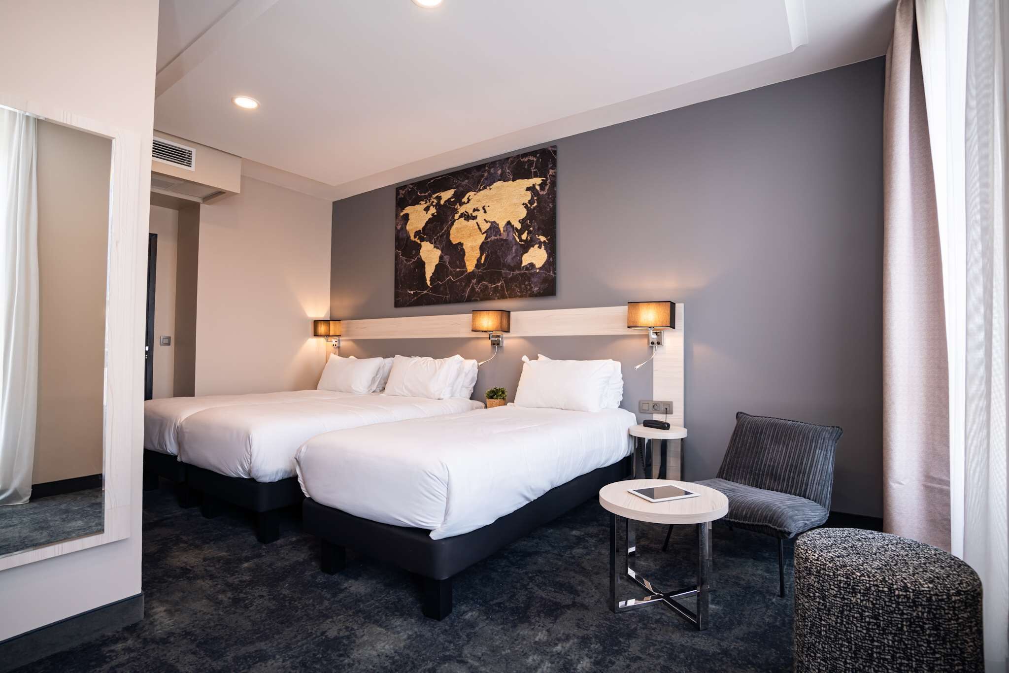 A hotel room with two beds, a small round table, a chair, and a world map artwork on the wall.