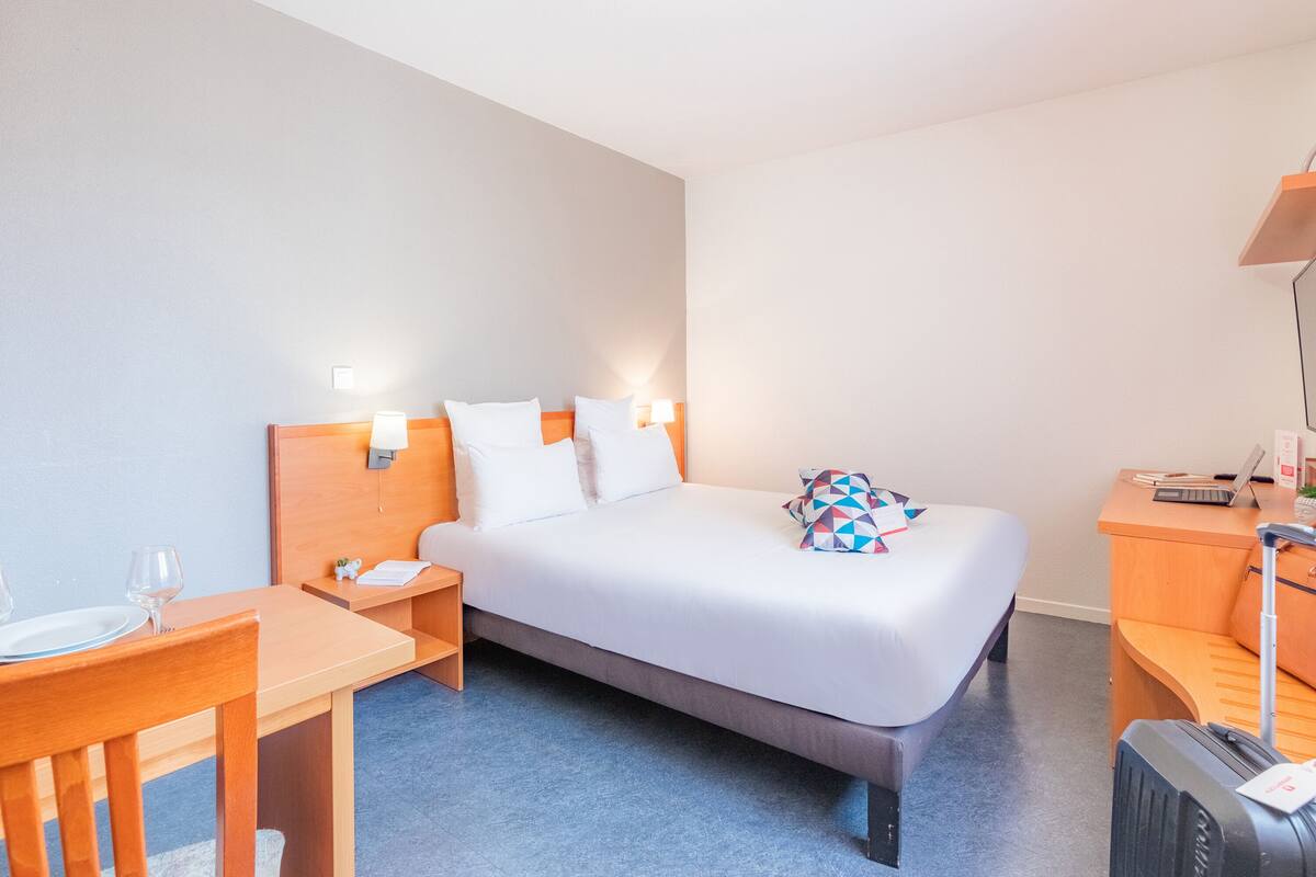 A hotel room with a bed, a desk, a chair, a table, and a suitcase.