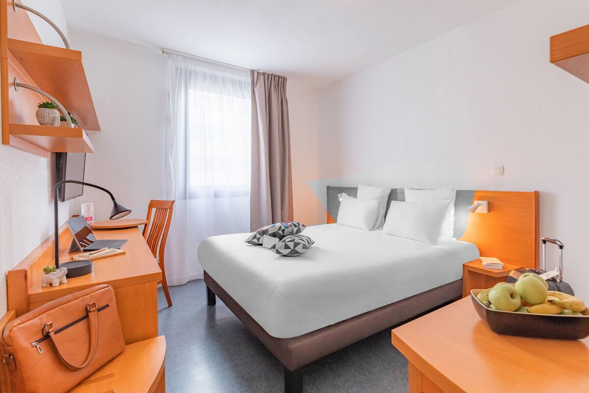 A hotel room with a bed, desk, chair, and a small table with fruit.