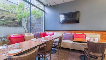 A modern dining area with a long table, cushioned seating, and a large window offering a view of trees and buildings.