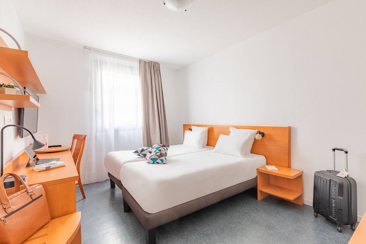A hotel room with a bed, a desk with a laptop, a chair, a nightstand, and a suitcase.