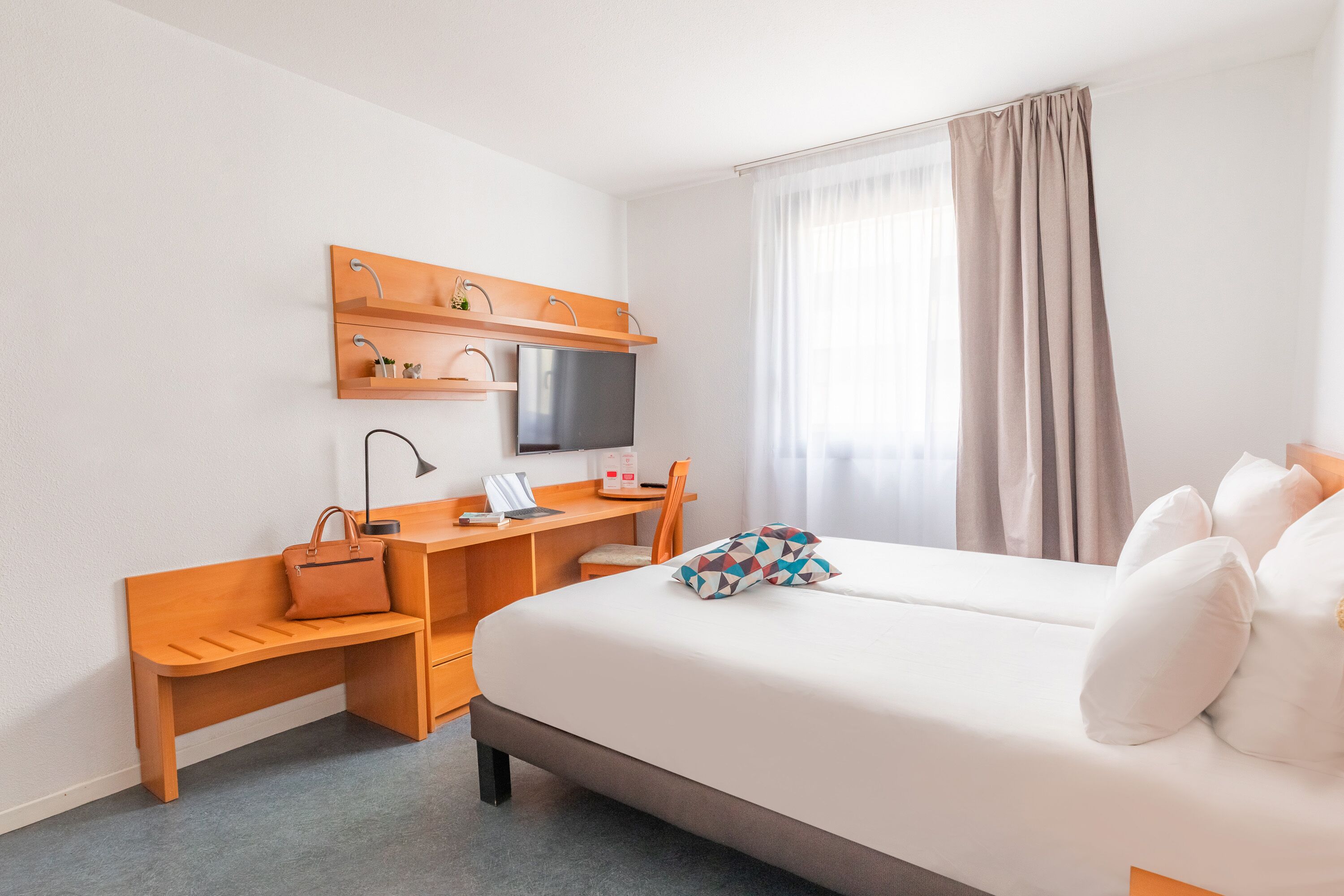 Una habitación de hotel con cama, escritorio, silla, televisión y ventana con cortinas.
