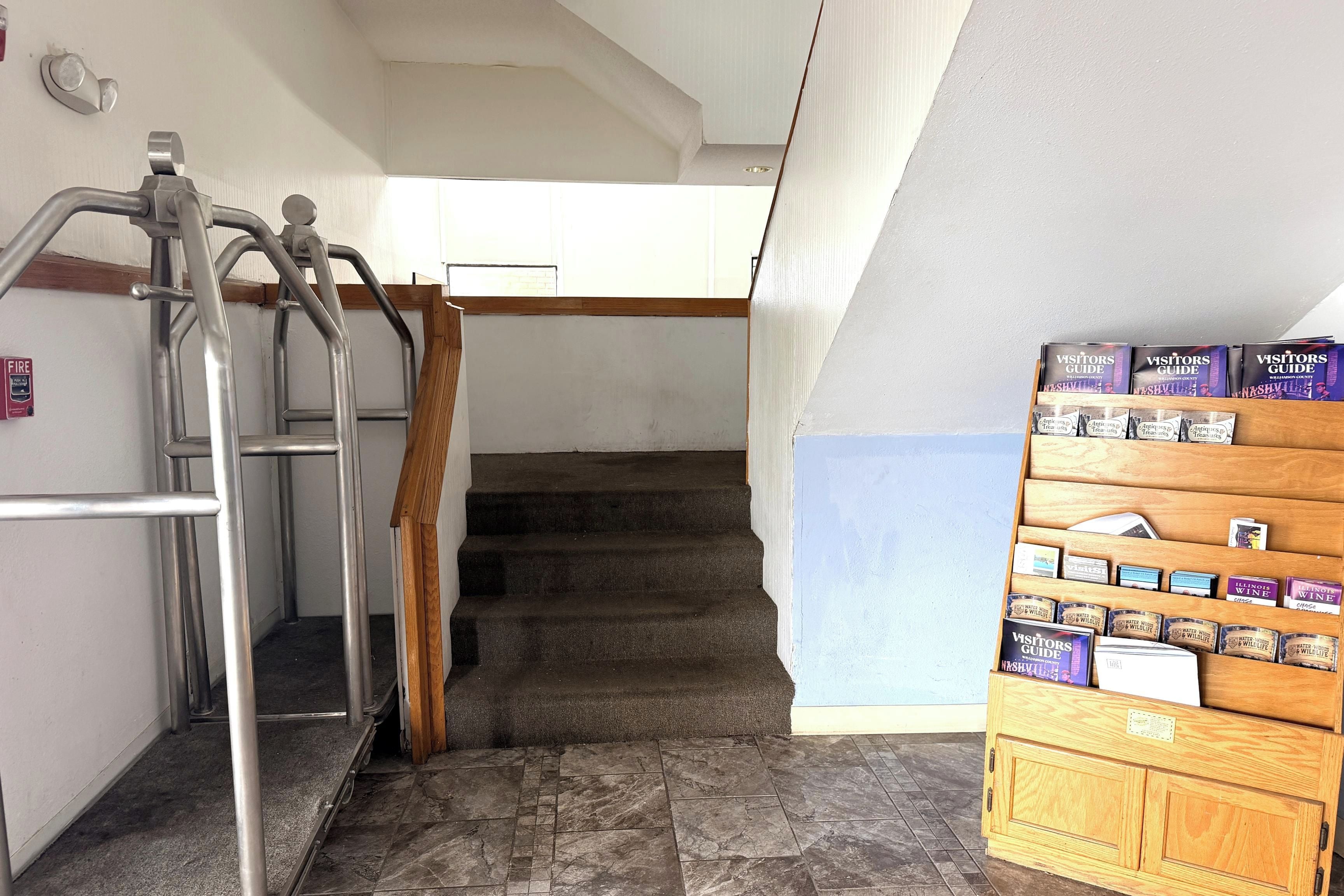 Una escalera con barandillas metálicas, un detector de incendios y un expositor de madera con guías para visitantes y cajas de vino.