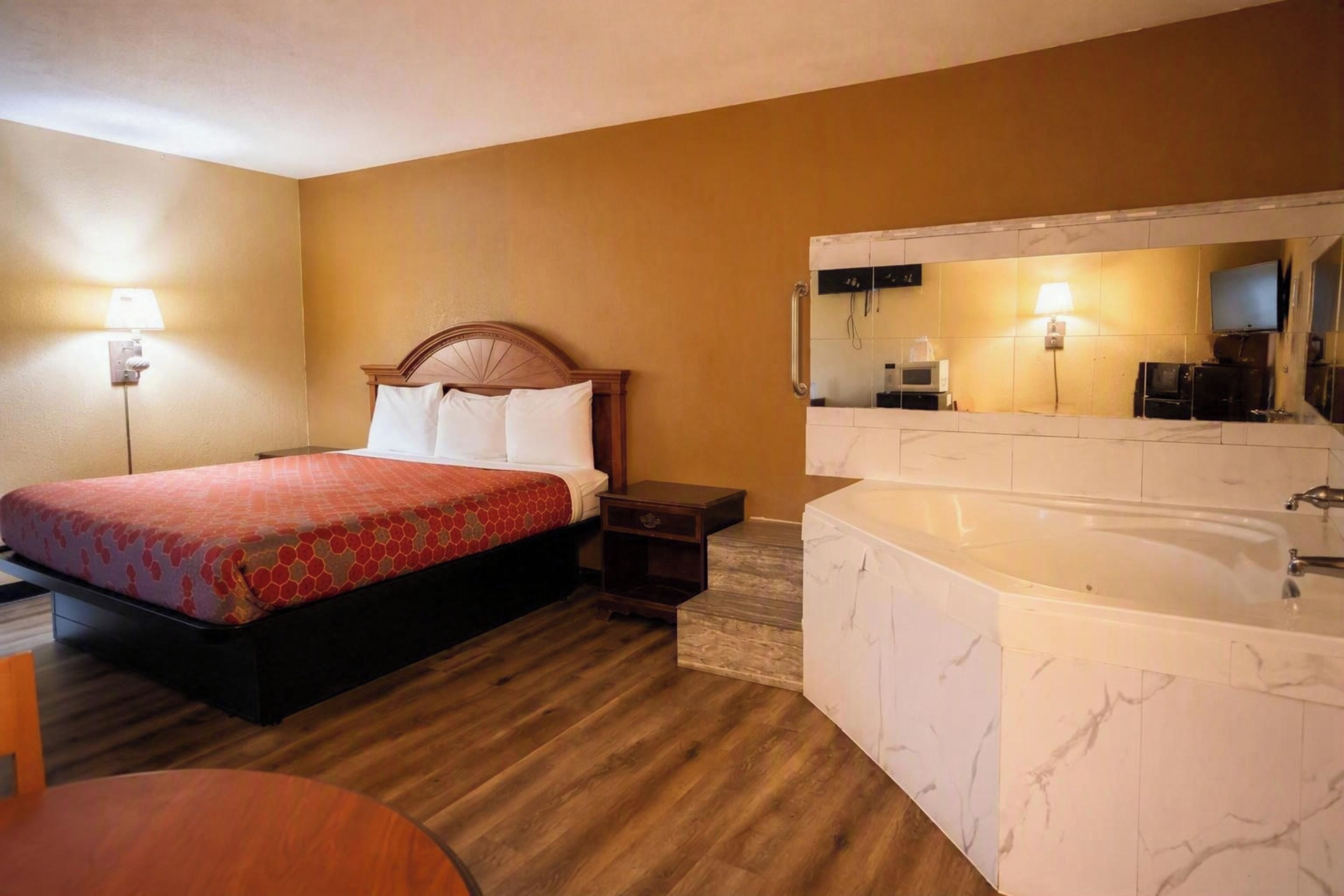 A hotel room with a bed, a nightstand, a desk, a chair, a large mirror, and a bathtub.