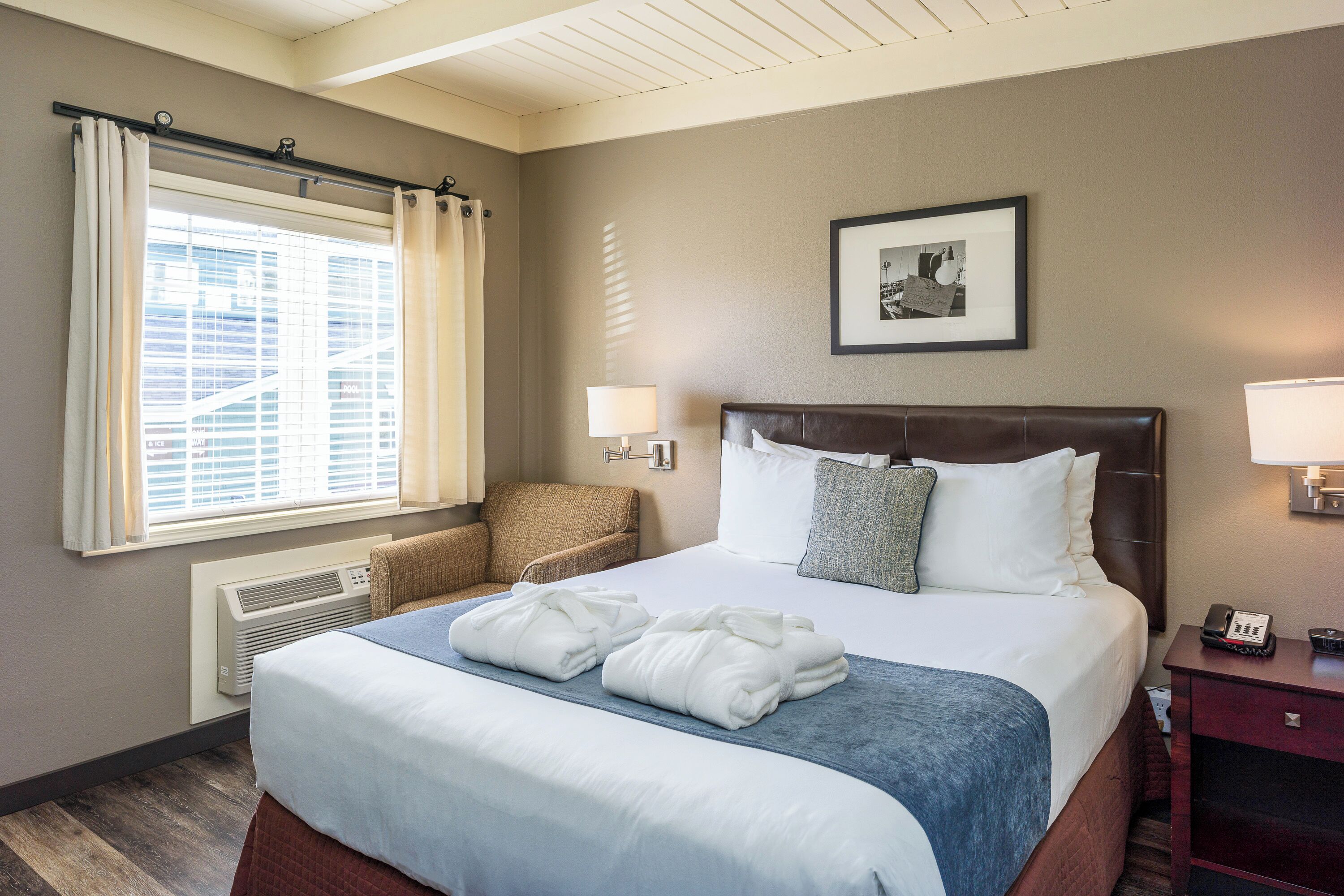 A hotel room with a large bed, a chair, a heater, a window with curtains, a framed picture on the wall, and a nightstand with a phone.
