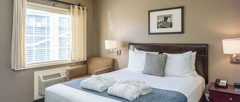 A hotel room with a large bed, a chair, a heater, a window with curtains, a framed picture on the wall, and a nightstand with a phone.