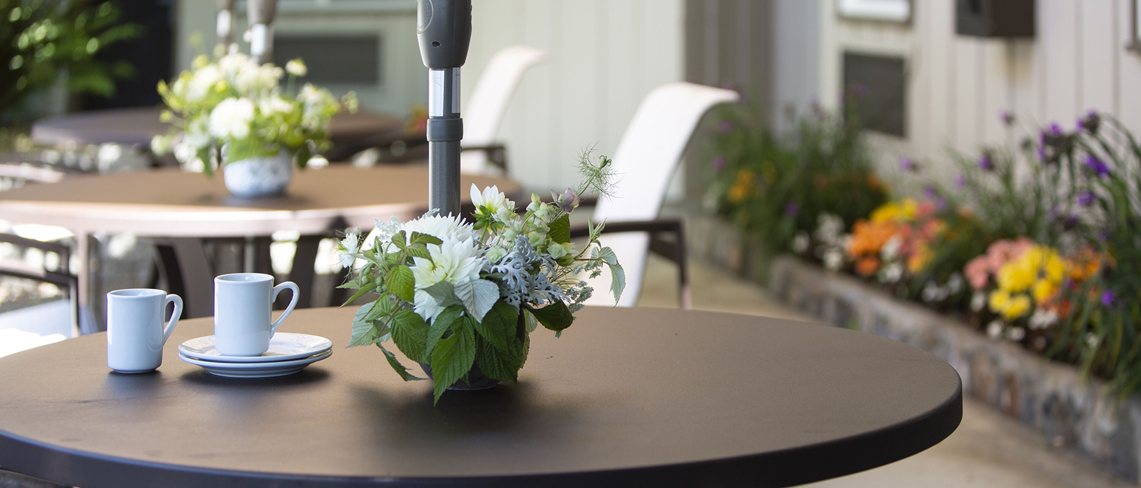 Ein runder Tisch mit einer weißen Tasse und Untertasse, einer Vase mit weißen Blumen und einer Pflanze im Hintergrund.