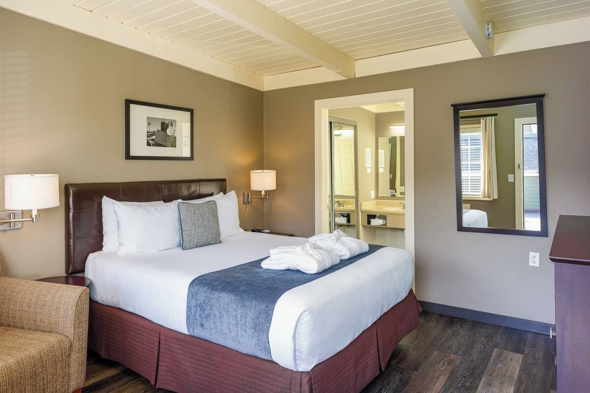 A hotel room with a large bed, a chair, a framed picture on the wall, and a mirror.