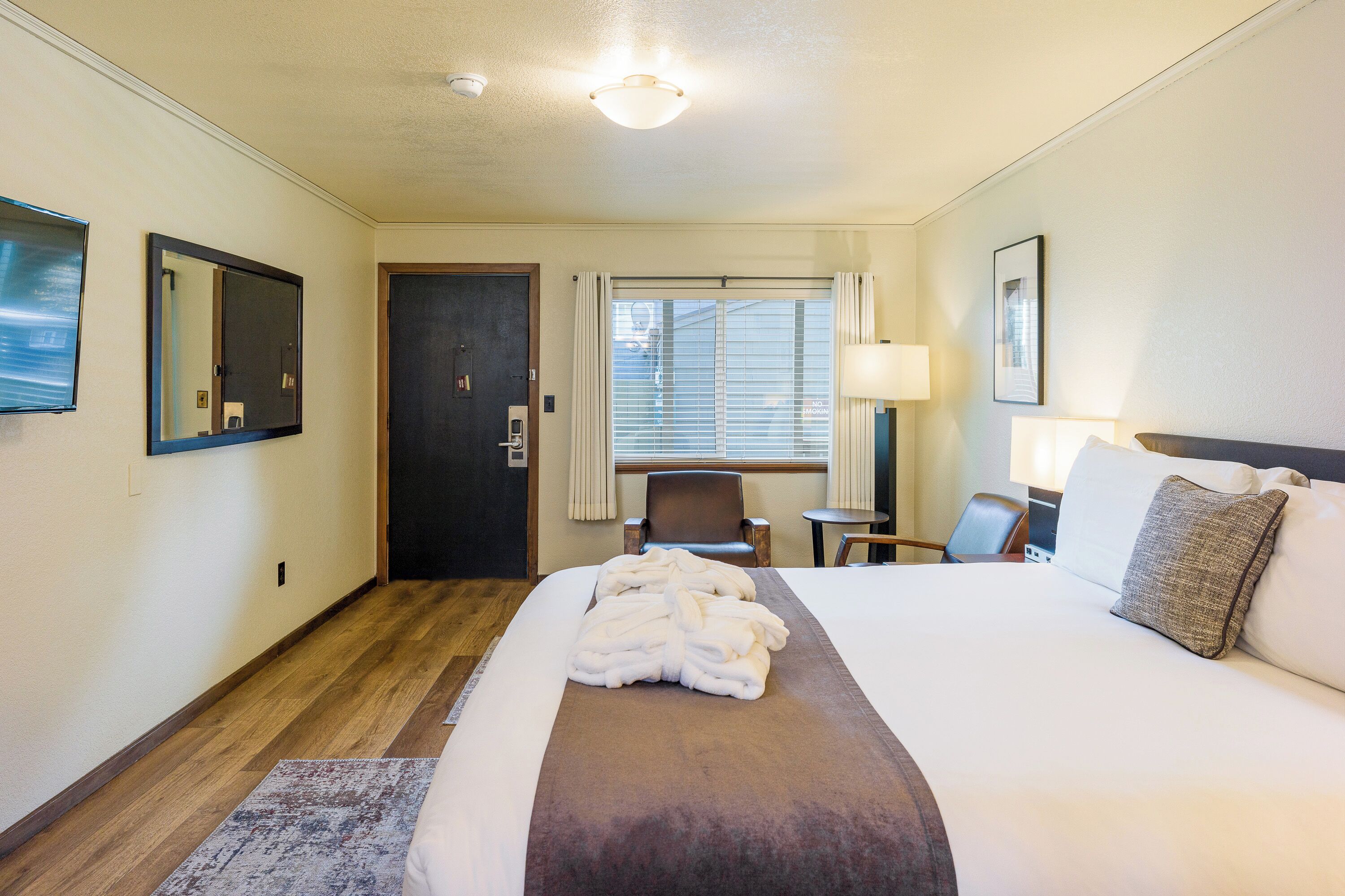 A hotel room with a large bed, a desk, a chair, a window with curtains, and a door.