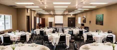 Une salle de conférence dotée de plusieurs tables rondes, préparées pour un repas, avec des nappes blanches, des chaises et des couverts.