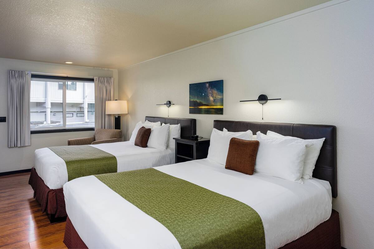 A hotel room with two beds, a large window with curtains, a nightstand, a lamp, and a wall-mounted clock.