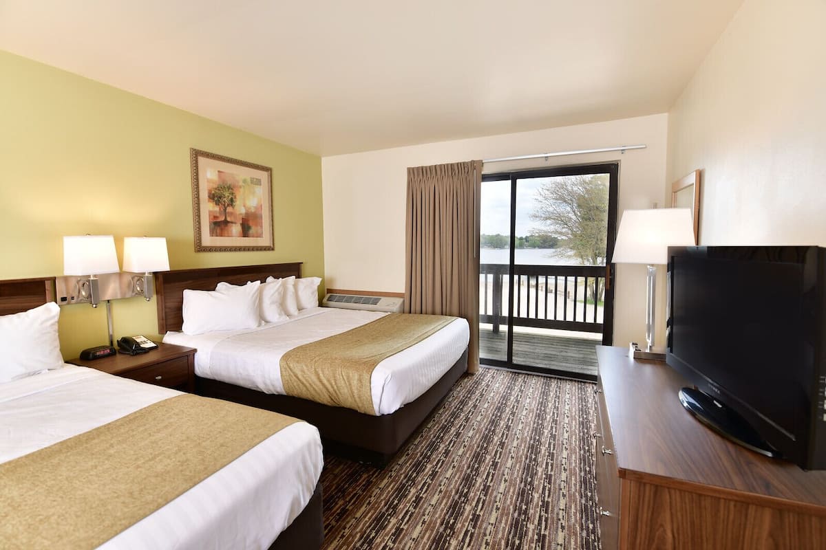 A hotel room with two beds, a TV, a desk, and a balcony with a view of the water.