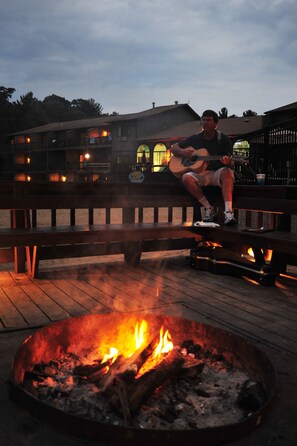 A person sitting on a bench near a fire pit with a building in the background. - Sunset Bay Resort & Suites (Wisconsin Dells)