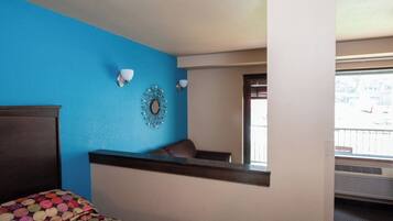 A hotel room with a blue accent wall, a bed with a colorful bedspread, a small seating area with a chair, and a decorative mirror on the wall.