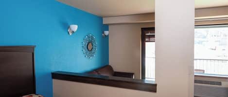 A hotel room with a blue accent wall, a bed with a colorful bedspread, a small seating area with a chair, and a decorative mirror on the wall.