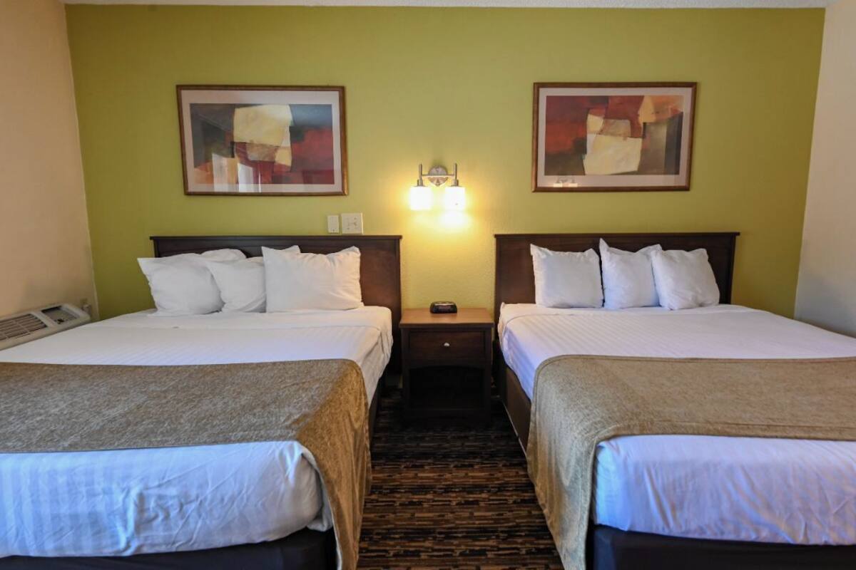 A hotel room with two beds, a headboard, a nightstand, a lamp, and two framed pictures on the wall.