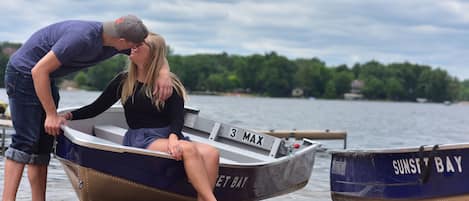 A couple kissing in a small boat on a sandy shore with another boat labeled "SUNSET BAY" in the background.