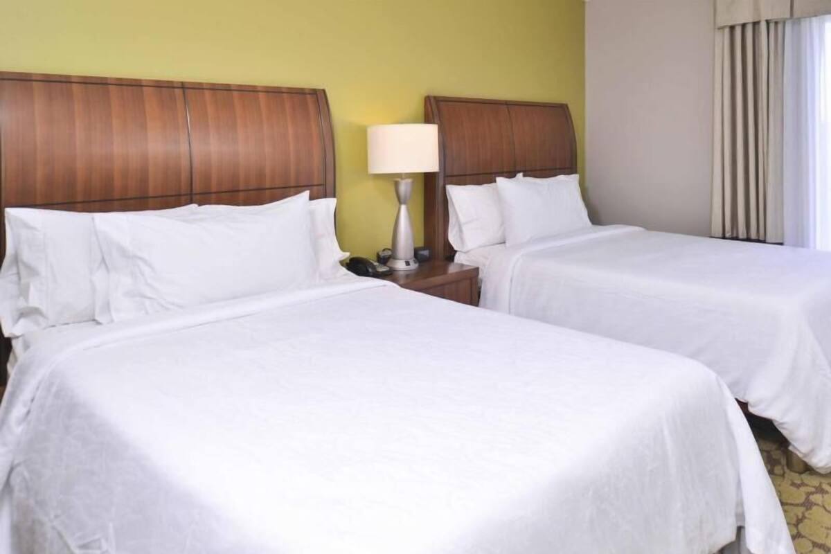 A hotel room with two beds, a nightstand with a lamp, and a window with curtains.