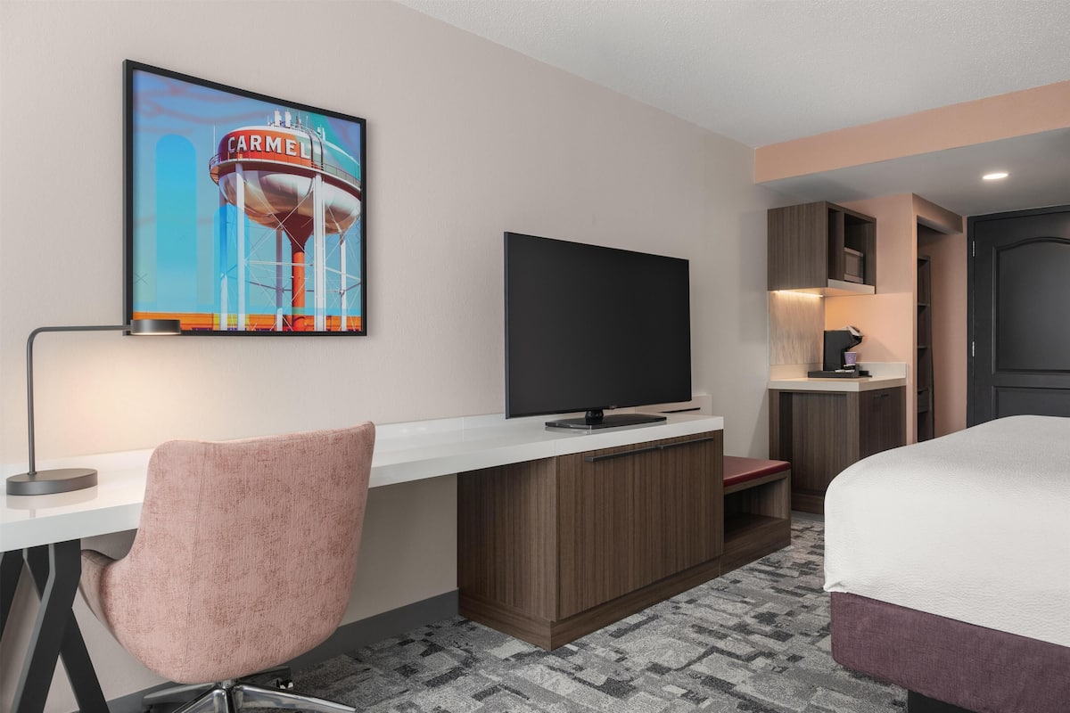 A hotel room with a bed, a desk with a television, a chair, a painting, and a small kitchenette.